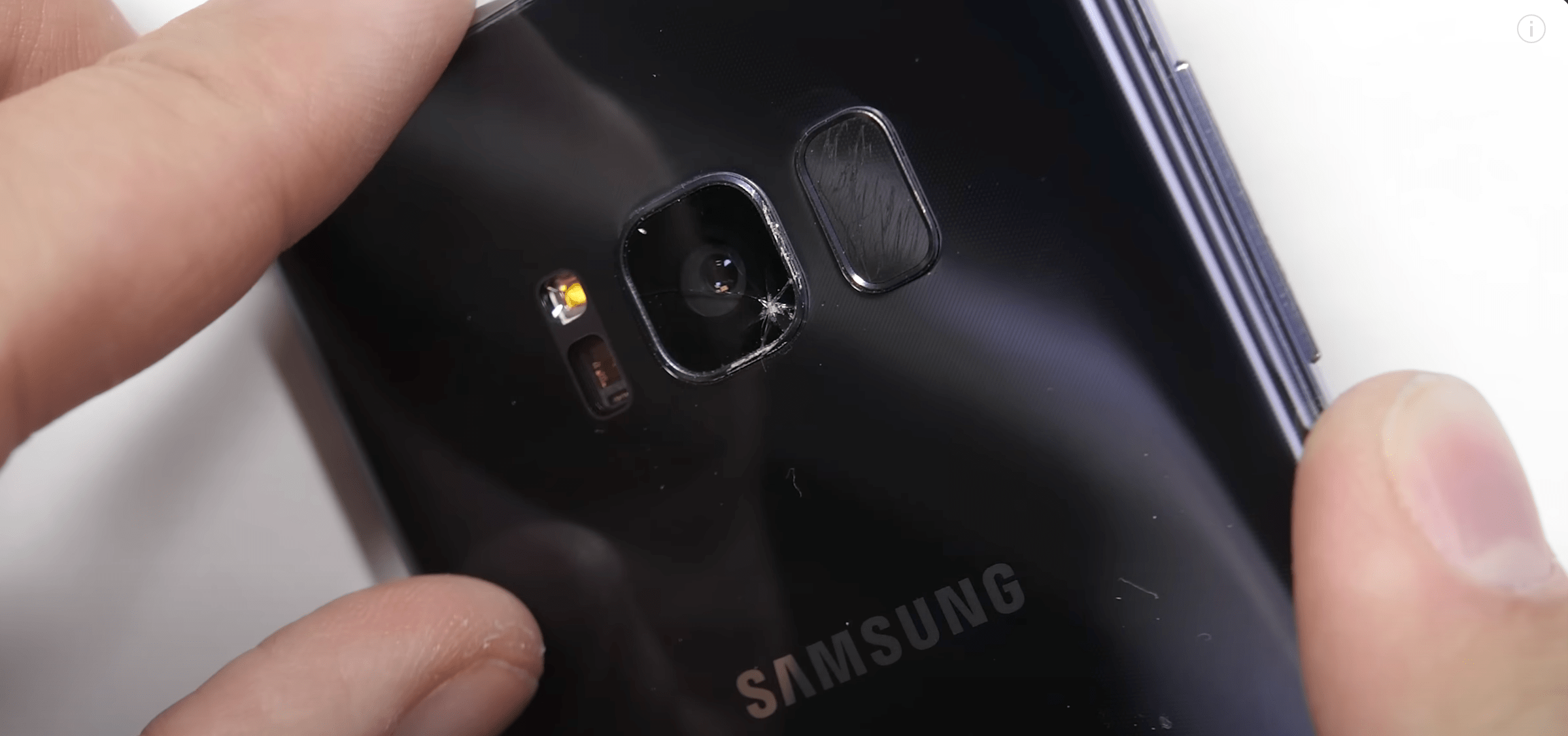 Close-up of a black Samsung smartphone's rear camera with a visible crack on the lens. A person's fingers hold the phone, suggesting concern or inspection.